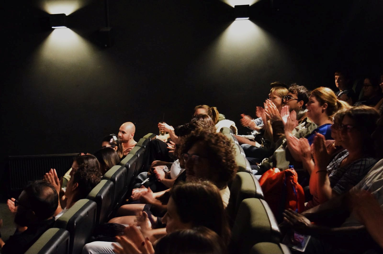 A full row of seats in a dark cinema, the audience applauding as the film ends.